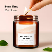 Load image into Gallery viewer, Candle labeled 'Reading by the Fire' being lit with a match on a light pink background.