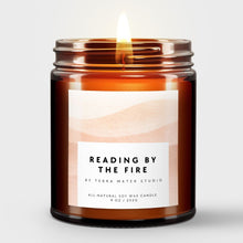 Load image into Gallery viewer, Scented candle in an amber jar with a label reading 'Reading by the Fire' by Terra Mater Studio on a light background.